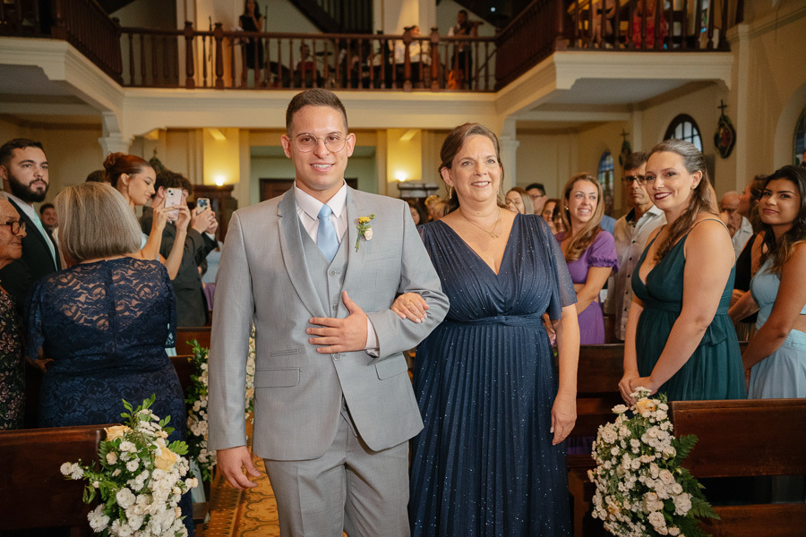 Entrada emocionante do noivo Marcus com sua mãe na cerimônia de casamento. Ele veste terno cinza claro e caminha pelo corredor da Capela no Parque Vicentina Aranha, cercado por convidados. O início solene do grande dia.
