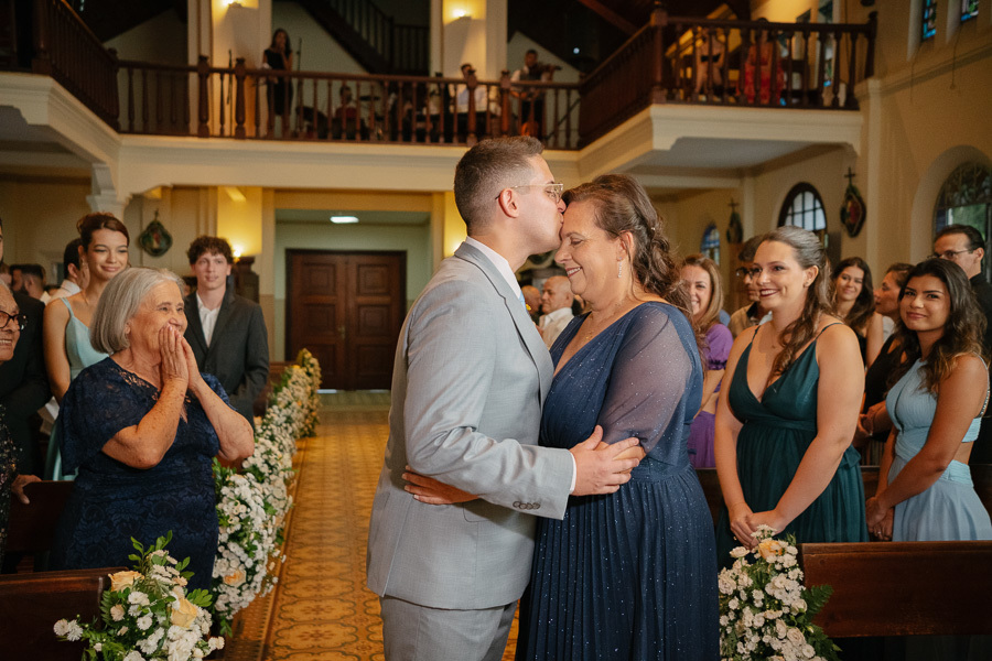 Noivo Marcus beija carinhosamente a testa de sua mãe ao chegar ao altar. Um momento de pura emoção e gratidão capturado na Capela Sagrado Coração de Jesus, com reações espontâneas da família ao fundo testemunhando o amor.