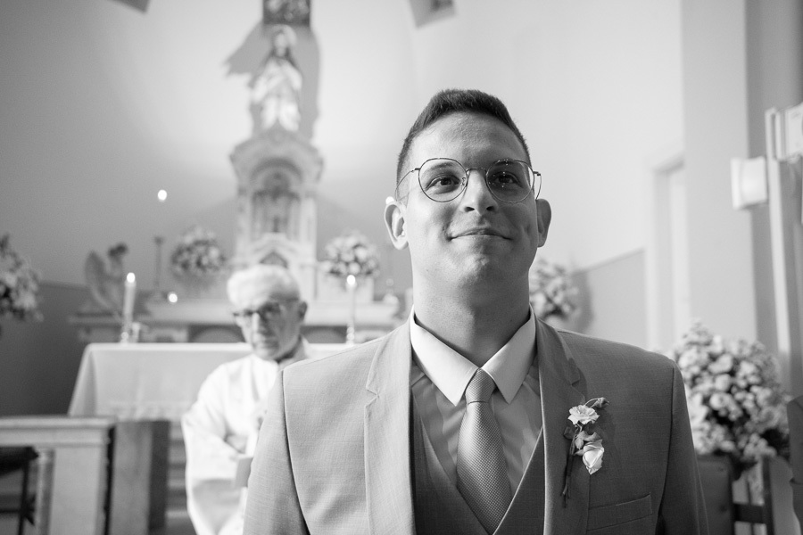 Noivo Marcus sorrindo emocionado no altar, aguardando a entrada da noiva. Retrato expressivo em preto e branco que destaca a ansiedade e a felicidade genuína do momento. Cerimônia religiosa clássica na Capela Sagrado Coração de Jesus.