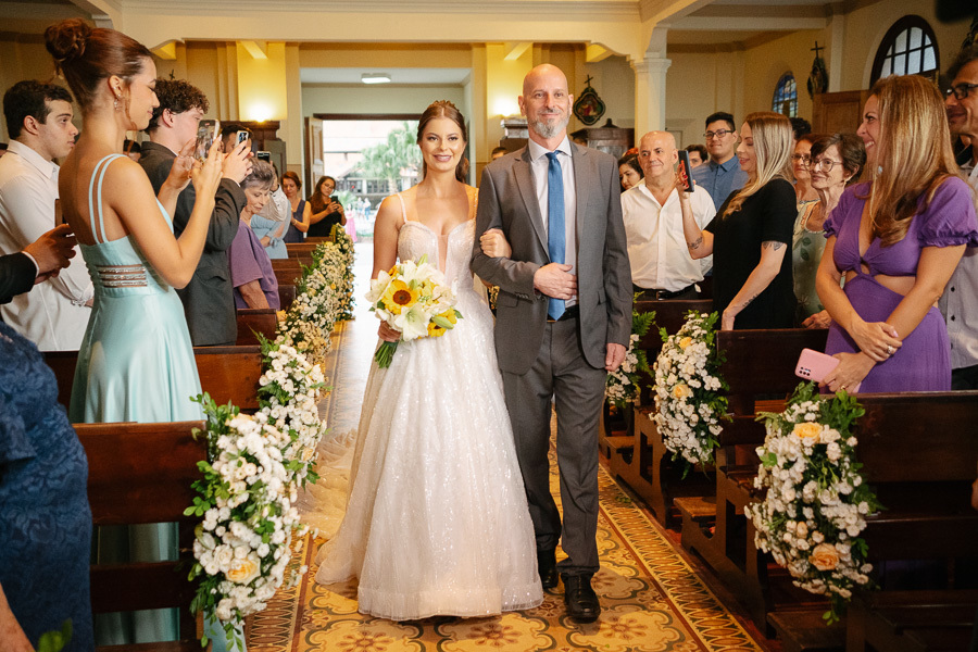 Entrada triunfal da noiva Isabelle caminhando pelo corredor da nave de braços dados com seu pai. Fotografia ampla em preto e branco capturando a reação dos convidados e a atmosfera solene na Capela Sagrado Coração de Jesus durante o cortejo.