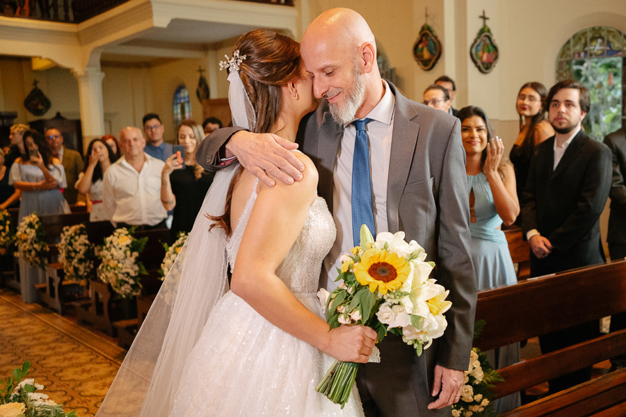Pai da noiva beija carinhosamente Isabelle ao entregá-la no altar. Um gesto emocionante de bênção paterna e despedida na Capela Sagrado Coração de Jesus. Fotografia que registra a forte conexão familiar no casamento.