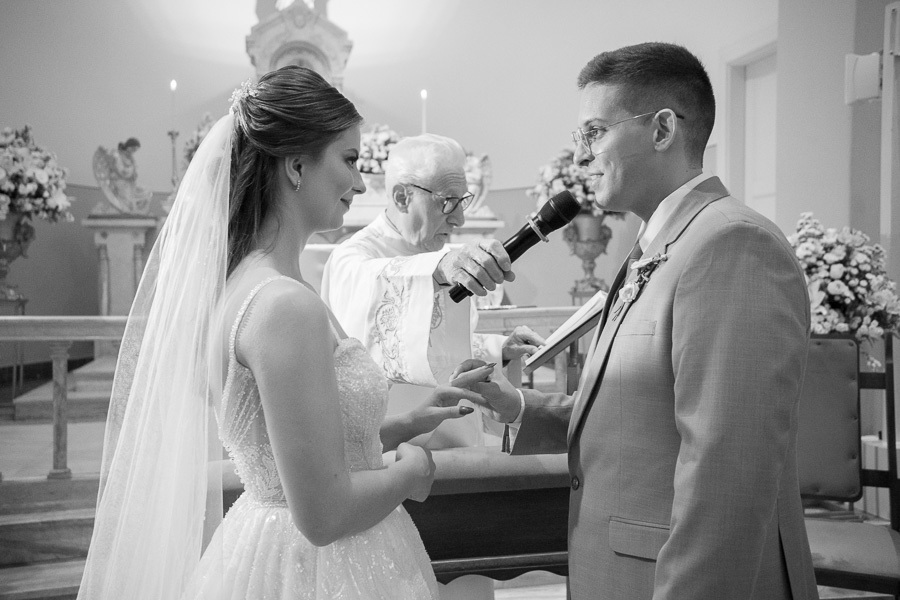 Momento solene da troca de alianças em preto e branco. O noivo Marcus coloca o anel em Isabelle sob a bênção do padre. Registro clássico e atemporal do compromisso selado na Capela Sagrado Coração de Jesus.