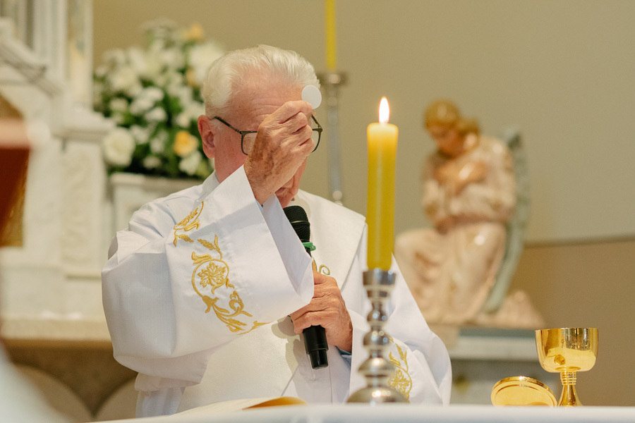 Sacerdote elevando a hóstia durante a consagração na cerimônia de casamento. Detalhe da liturgia católica na Capela Sagrado Coração de Jesus, trazendo a sacralidade e a tradição religiosa para a narrativa do dia.