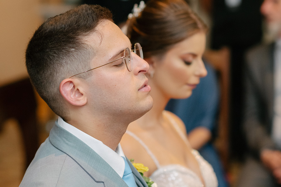 Noivo Marcus de olhos fechados em momento de profunda oração e gratidão ao lado da noiva. Fotografia de perfil que destaca a espiritualidade e a emoção intensa vivida no altar durante o casamento religioso.