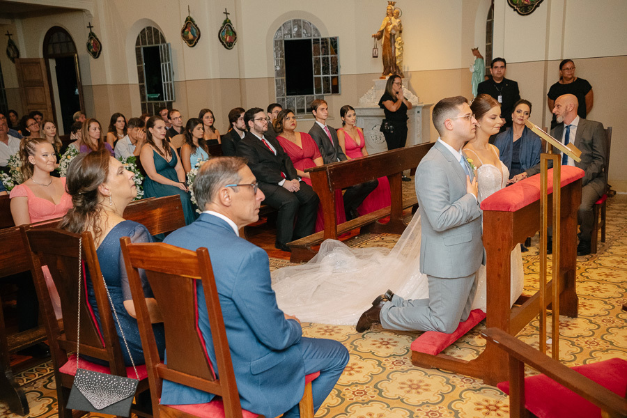 Marcus e Isabelle ajoelhados recebendo a bênção final no altar da Capela Sagrado Coração de Jesus. Um momento de fé e conexão familiar, com os pais emocionados ao fundo. Fotografia respeitosa de cerimônia religiosa no Parque Vicentina Aranha.