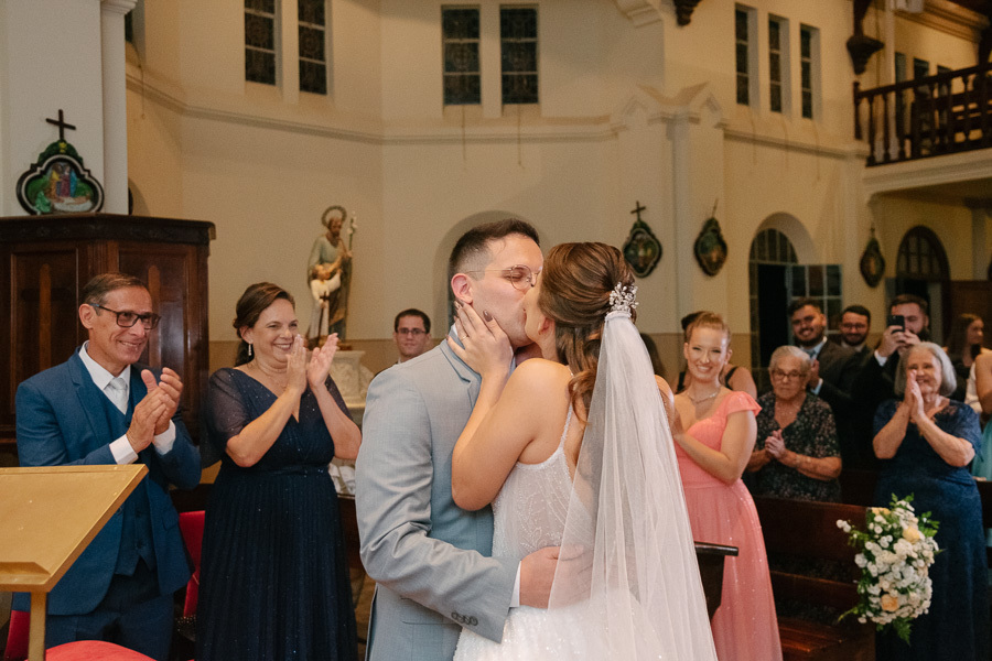 O primeiro beijo de Marcus e Isabelle como marido e mulher no altar. Fotografia ampla mostrando a arquitetura sacra e a decoração floral da Capela Sagrado Coração de Jesus. O clímax romântico da celebração de casamento.