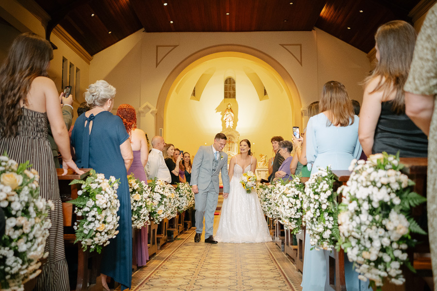 Recém-casados Marcus e Isabelle caminhando sorridentes pelo corredor da nave após o 'sim'. Saída dos noivos na Capela Sagrado Coração de Jesus cercados pelo carinho dos convidados. O início da nova vida a dois.