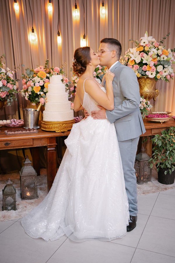 O beijo apaixonado dos noivos na mesa do bolo durante a recepção na APVE Embraer. Decoração rústica e romântica com girassóis, luzes pendentes e bolo de cinco andares. O início vibrante da festa de casamento.
