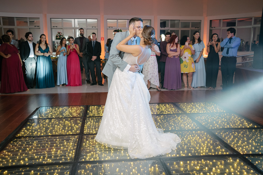 Noivos protagonizam um beijo de cinema na pista de dança. Marcus inclina Isabelle em um gesto romântico cercado pelos convidados na festa. Fotografia espontânea que captura o amor e a animação do casamento na APVE Embraer.