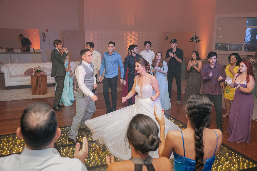 Noiva Isabelle girando seu vestido de princesa na pista de LED enquanto o noivo Marcus dança animado. A festa de casamento na APVE Embraer em seu auge, com convidados ao redor vibrando com a energia do casal.