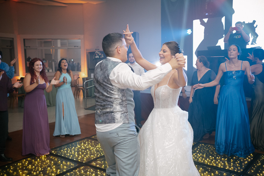 Marcus e Isabelle sorrindo enquanto dançam de mãos dadas sob a iluminação cênica. Um momento de diversão e cumplicidade dos noivos na pista de dança, celebrando o amor com movimentos leves e espontâneos.