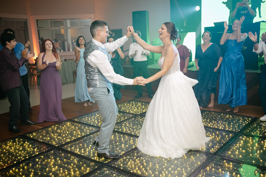 A alegria contagiante de Marcus e Isabelle dançando frente a frente na recepção do casamento. Sorrisos largos e olhares trocados na pista da APVE Embraer, cercados por amigos e familiares aplaudindo.