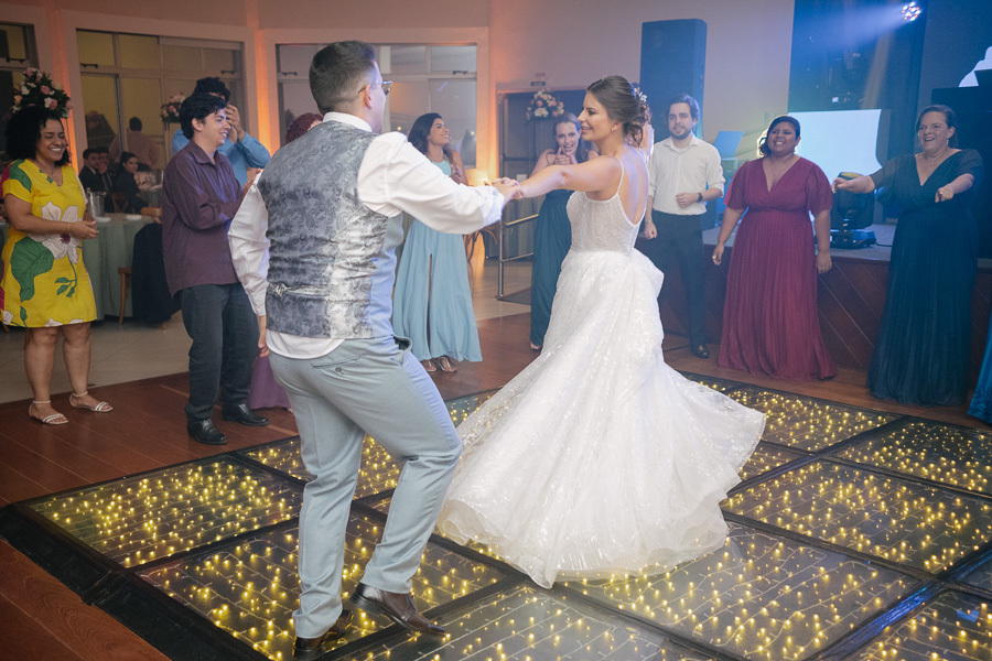 Noiva Isabelle gargalhando espontaneamente na pista de dança. Retrato de felicidade genuína iluminado pelas luzes da festa na APVE Embraer. O registro perfeito de uma noiva radiante aproveitando cada segundo do seu casamento.