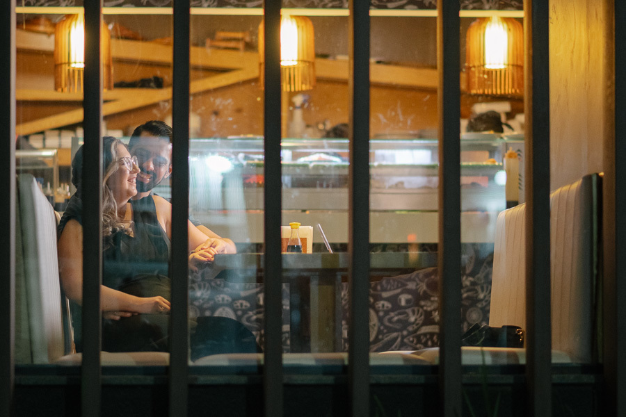 Ensaio pré-casamento de Tainara e Juliano no Hioki Sushi em São José dos Campos. Foto lifestyle através do vidro, capturando intimidade. Fotógrafo de casamento em SJC e Vale do Paraíba especializado em ensaios de casal espontâneos e urbanos.
