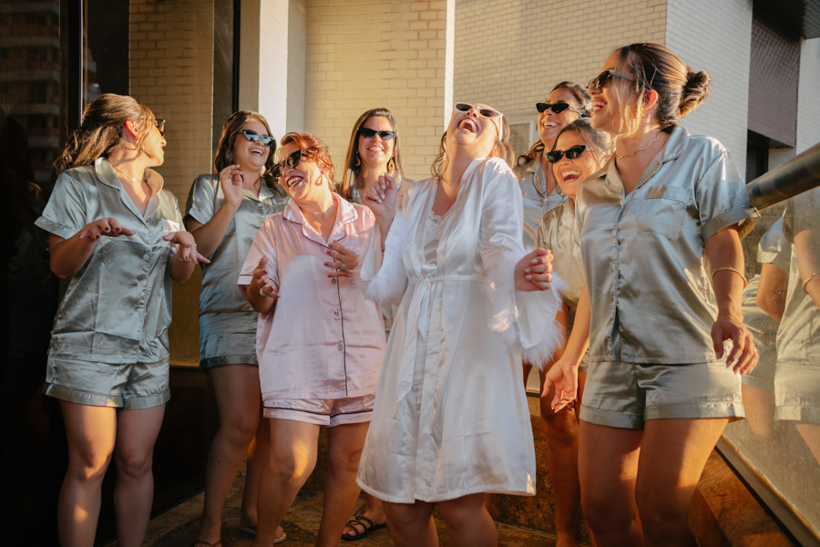 Noiva Gabriela e madrinhas rindo e dançando na varanda. Fotografia espontânea do making of capturando a alegria contagiante e a conexão entre amigas. Preparativos vibrantes para um casamento inesquecível.