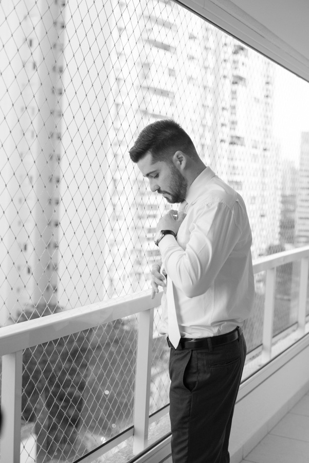 Making of do noivo Lucas em preto e branco. Retrato concentrado ajustando a gravata próximo à janela com luz natural. Preparação masculina clássica e elegante antes da cerimônia na Capela Nossa Senhora de Fátima.