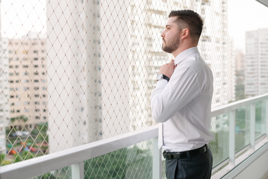 Noivo Lucas ajustando a gravata na varanda com luz natural. Retrato clássico e sereno da preparação masculina antes da cerimônia. Ele veste camisa branca e observa o horizonte, pronto para o grande dia.