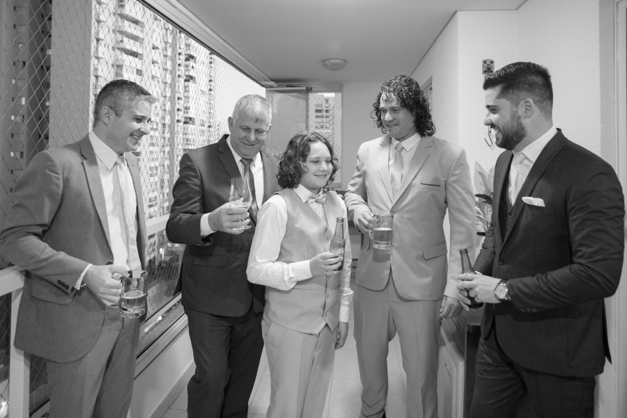 Making of descontraído do noivo Lucas e padrinhos brindando com cerveja na varanda. Fotografia em preto e branco capturando risadas e a camaradagem masculina antes da cerimônia. Um momento leve e divertido entre amigos e o pajem.