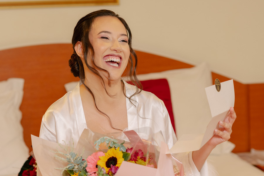 Noiva Gabriela gargalhando emocionada ao ler uma carta especial durante o making of. Fotografia espontânea que registra a felicidade genuína e a expectativa antes do casamento na Capela Nossa Senhora de Fátima.