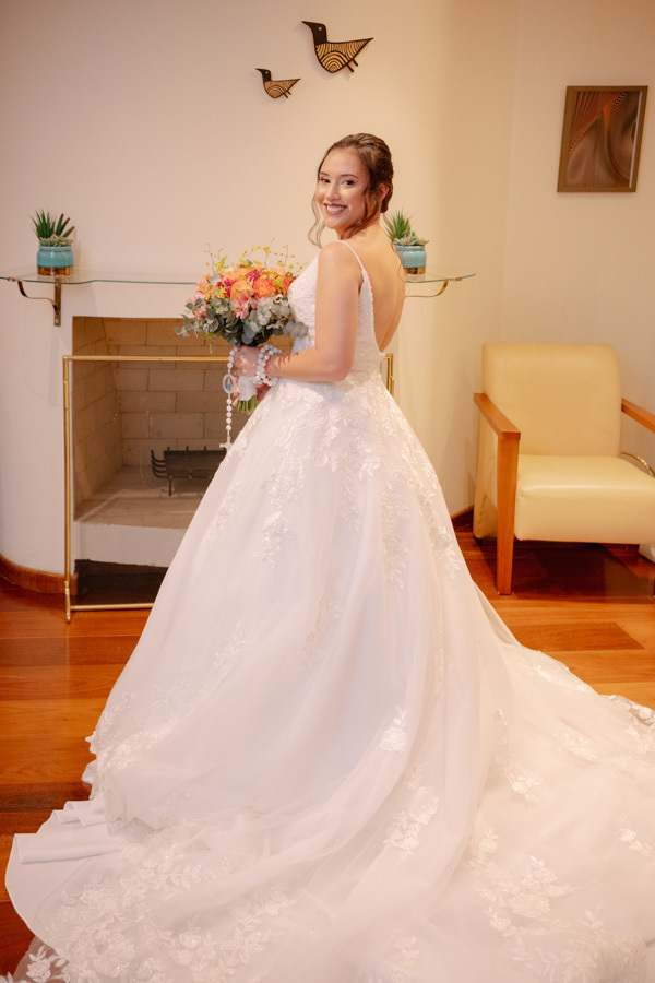 Retrato da noiva Gabriela pronta, exibindo o decote nas costas do vestido de noiva e seu buquê colorido. Pose elegante que destaca os detalhes da renda e a felicidade radiante antes de sair para o casamento.