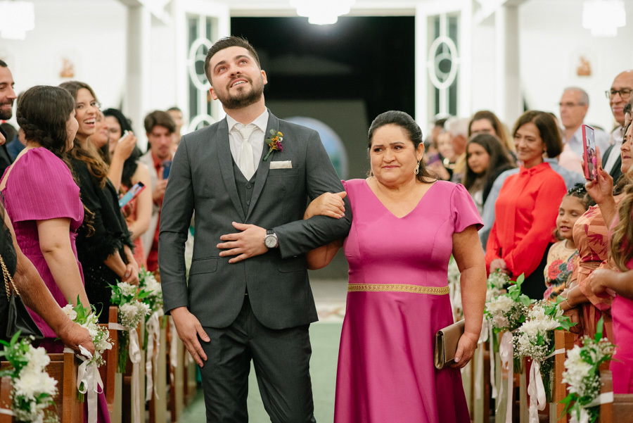 Entrada emocionante do noivo Lucas com sua mãe na igreja. Ele olha para o alto, contendo a emoção a caminho do altar. Cerimônia de casamento clássica e tocante realizada na Capela Nossa Senhora de Fátima.