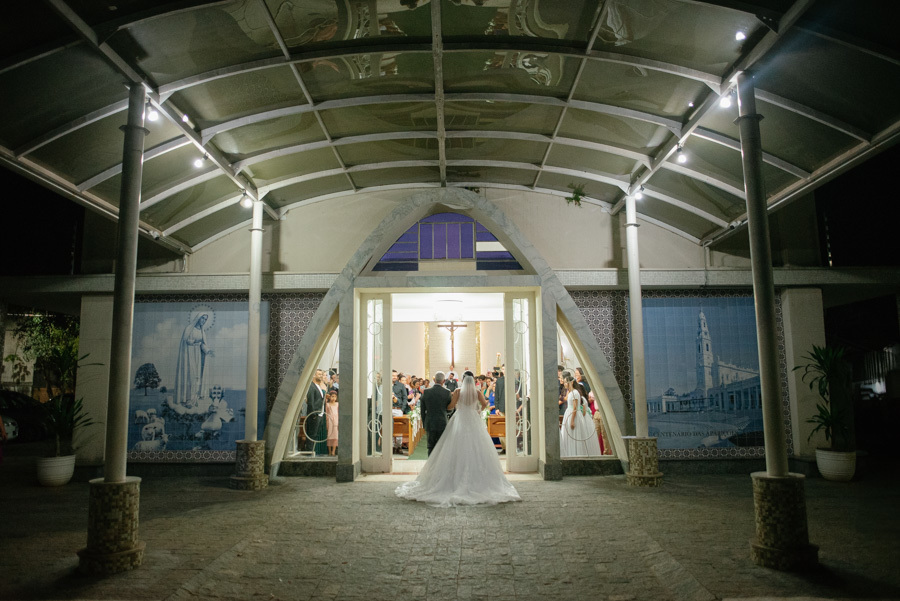 Vista ampla e noturna da entrada da noiva Gabriela na Capela Nossa Senhora de Fátima. A arquitetura iluminada da igreja e a longa cauda do vestido criam uma cena mágica e solene para o início da cerimônia.