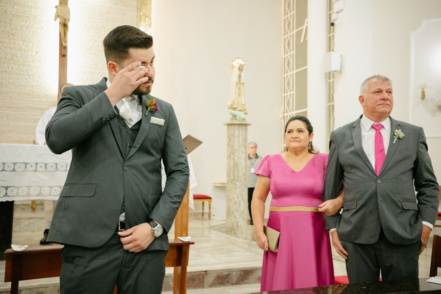 Noivo Lucas emocionado enxugando as lágrimas no altar ao ver a entrada da noiva. Um momento de pura sensibilidade e amor na cerimônia religiosa na Capela Nossa Senhora de Fátima. Reação espontânea e inesquecível do noivo.