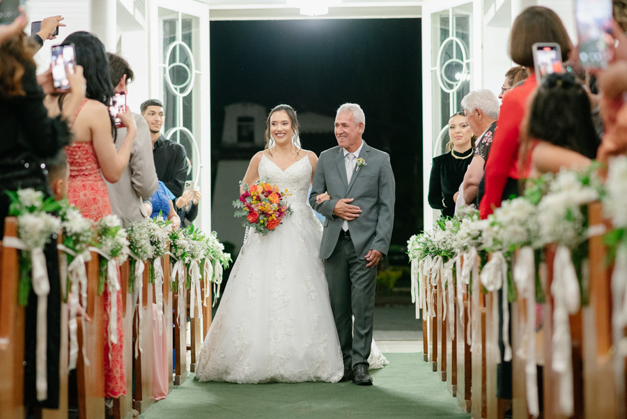 Entrada radiante da noiva Gabriela de braços dados com seu pai. Ela caminha sorrindo pelo corredor da Capela Nossa Senhora de Fátima, segurando seu buquê colorido. A alegria contagiante da noiva a caminho do altar.