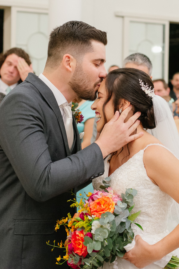 Lucas beija carinhosamente a testa de Gabriela no encontro no altar. O casal sorri demonstrando cumplicidade e felicidade. Destaque para as cores vibrantes do buquê da noiva contrastando com o terno cinza do noivo.
