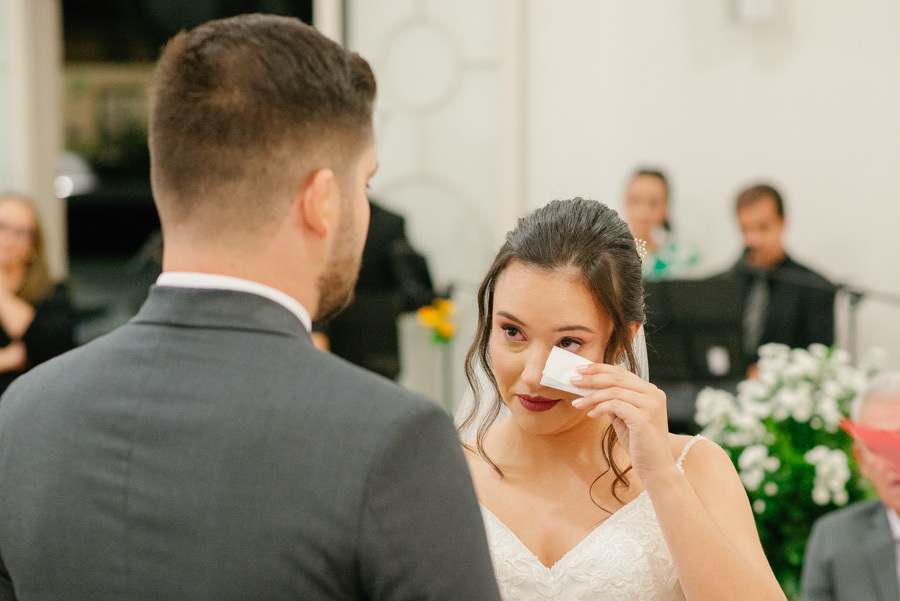 Close emocionante da noiva Gabriela enxugando as lágrimas ao olhar para Lucas. O registro autêntico de um momento de vulnerabilidade e felicidade extrema durante os votos na Capela Nossa Senhora de Fátima.