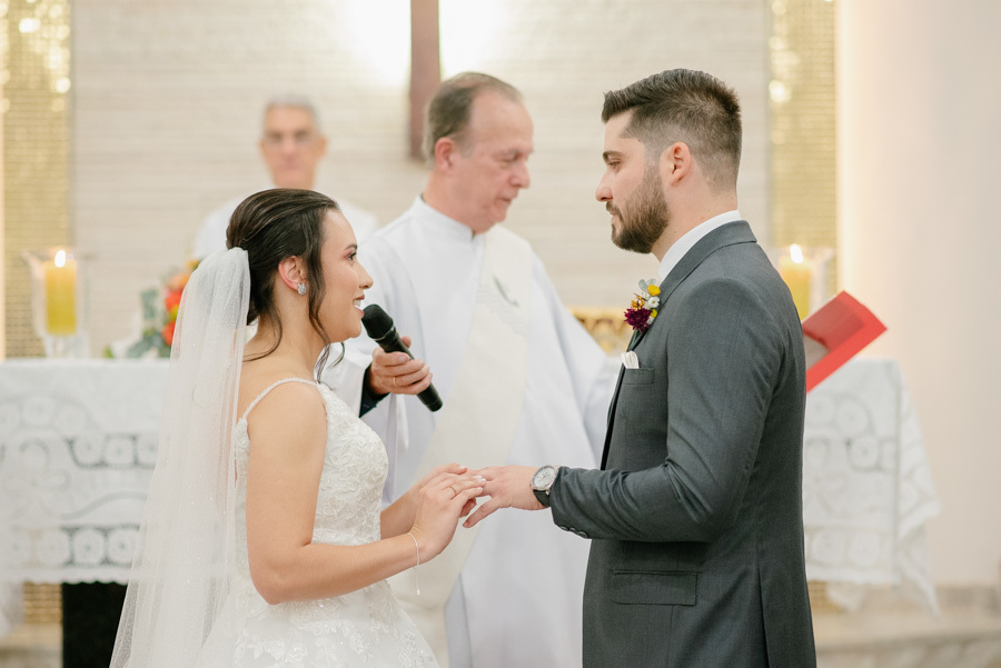 Momento sagrado da troca de alianças. Gabriela coloca a aliança no dedo de Lucas sob o olhar atento do padre. Detalhe clássico da união e do compromisso selado na cerimônia religiosa de casamento.
