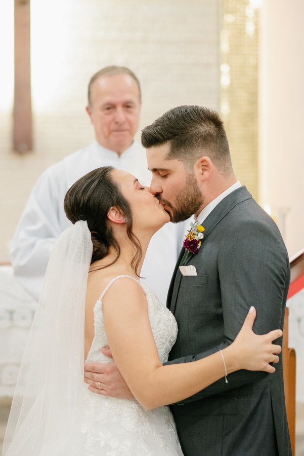 O beijo apaixonado de Lucas e Gabriela selando o casamento no altar. Momento romântico celebrado pelo padre na Capela Nossa Senhora de Fátima. O primeiro beijo como marido e mulher, marcando o início de uma nova vida.