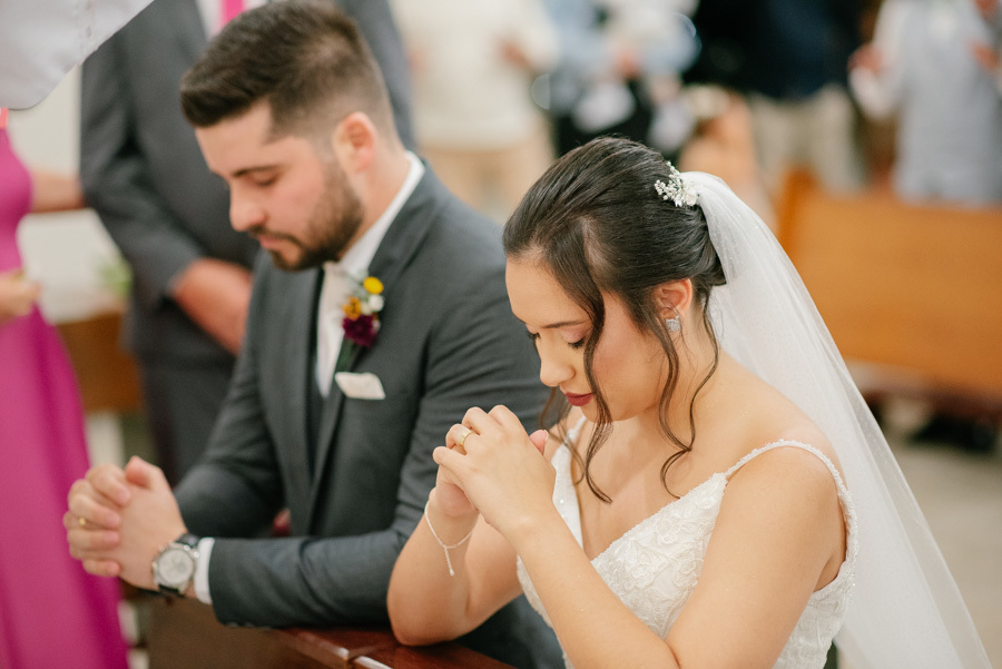 Lucas e Gabriela ajoelhados em oração durante a cerimônia religiosa. Um momento de fé e devoção na Capela Nossa Senhora de Fátima. O casal recebe a bênção final de olhos fechados, agradecendo pela união.