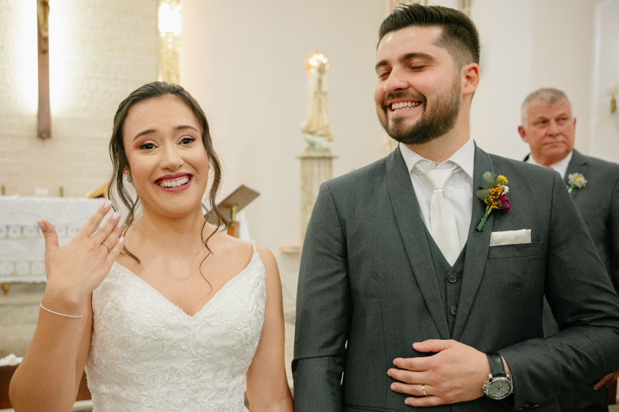 Noiva Gabriela exibe orgulhosa sua aliança de casamento enquanto Lucas ri espontaneamente ao lado. Um flagra divertido e cheio de personalidade no altar, quebra de protocolo que mostra a alegria genuína do casal.
