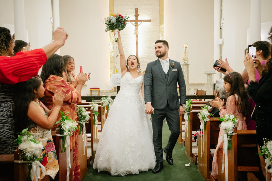Saída triunfal dos noivos com a noiva Gabriela erguendo o buquê em comemoração. Lucas e Gabriela caminham pelo corredor da igreja sob aplausos dos convidados. A vibração e a euforia do 'recém-casados' na Capela Nossa Senhora de Fátima.