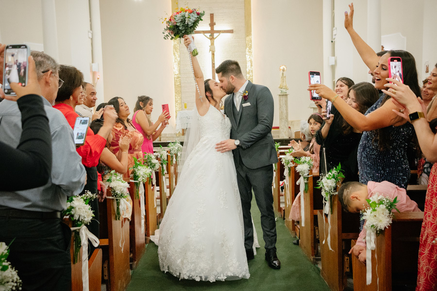 aída vibrante dos recém-casados Lucas e Gabriela na Capela Nossa Senhora de Fátima. A noiva ergue o buquê celebrando o amor enquanto caminham pelo corredor, cercados por convidados emocionados registrando o momento.