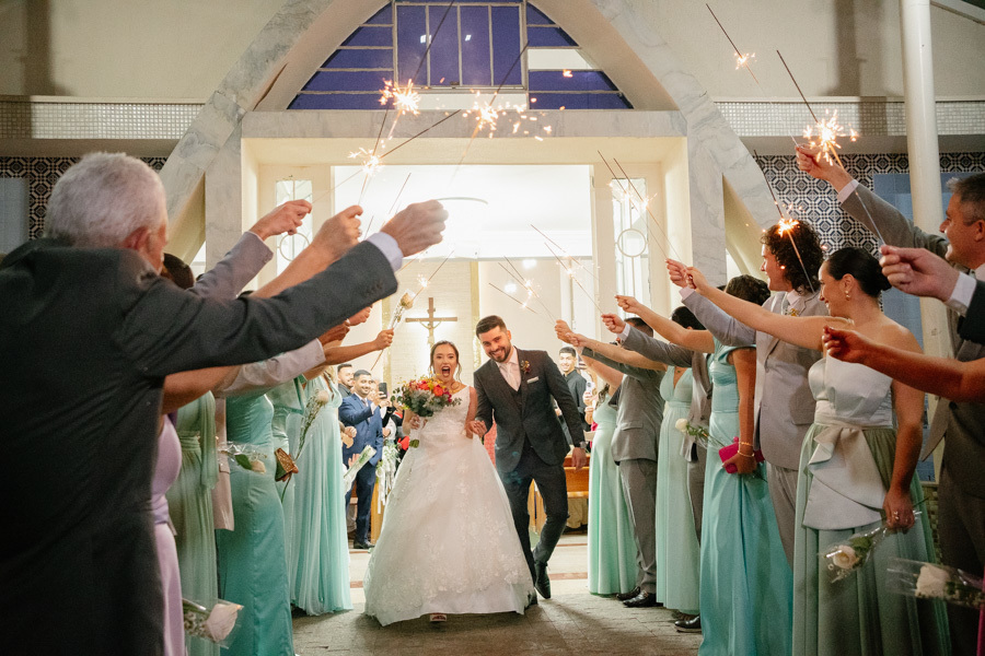aída mágica dos noivos com sparkles (bengalas de fogo). Lucas e Gabriela caminham sob um túnel de luzes segurado pelos padrinhos na saída da igreja. Fotografia noturna criativa e brilhante antes da festa.