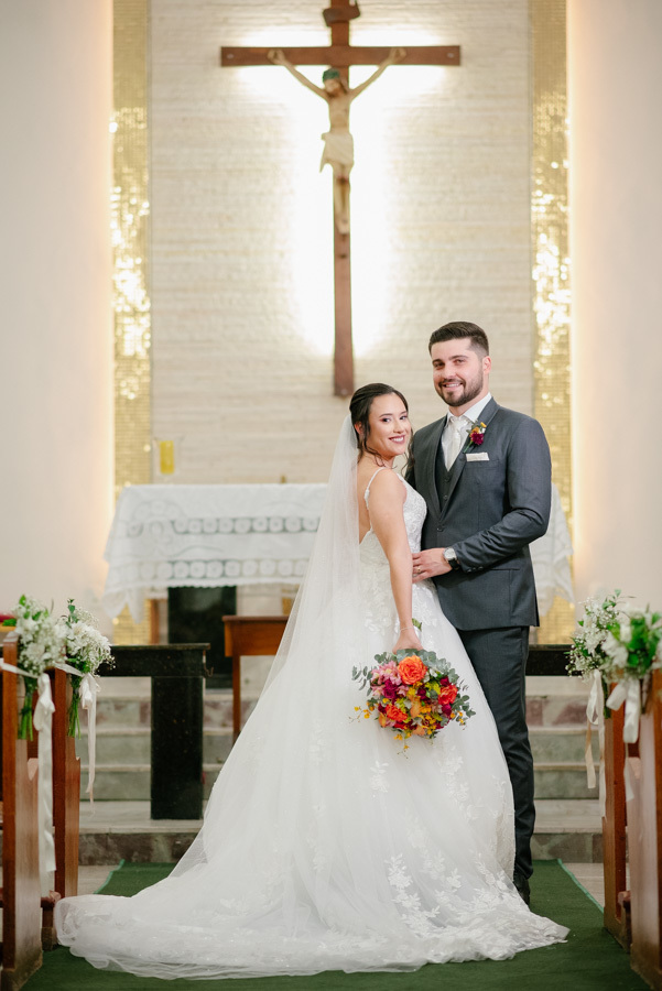 Retrato clássico de Lucas e Gabriela no altar da Capela Nossa Senhora de Fátima. O casal posa com elegância após o 'sim', com destaque para os detalhes do vestido de noiva e o buquê colorido. A foto oficial do casamento.
