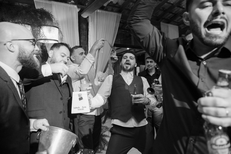 Animação total durante a hora da gravata com o noivo Lucas e seus amigos. Fotografia em preto e branco capturando a descontração e as brincadeiras na pista de dança da Quinta das Palmeiras. Tradição divertida para a lua de mel.