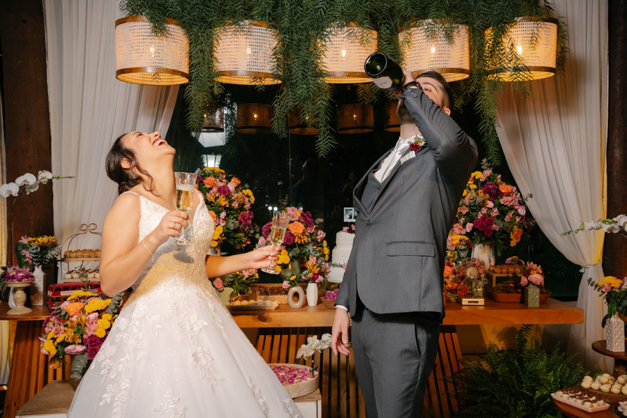 Lucas vira a garrafa de espumante enquanto Gabriela brinda rindo na mesa do bolo. Cena divertida e espontânea sob a decoração de luminárias de palha na Quinta das Palmeiras. A celebração eufórica dos recém-casados.