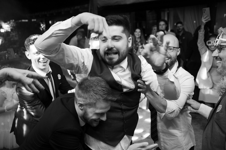 Noivo Lucas sendo jogado para o alto pelos padrinhos na pista de dança. O auge da festa de casamento registrado em preto e branco, transmitindo a euforia e a bagunça organizada dos amigos. Momentos inesquecíveis de pura energia.
