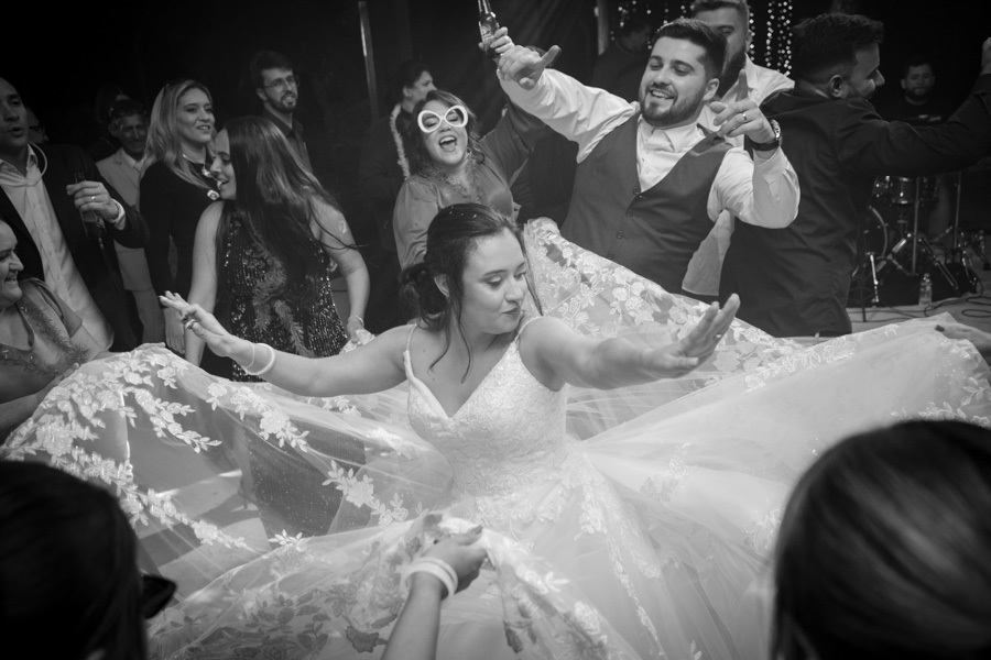 Gabriela e Lucas no centro da roda de dança cercados pelos amigos. Convidados usam adereços divertidos e óculos engraçados na pista. A energia contagiante e a bagunça boa da festa de casamento na Quinta das Palmeiras.