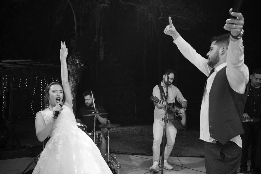 Lucas e Gabriela assumem o microfone e cantam com a banda ao vivo no palco. Noivos agitando a festa na Quinta das Palmeiras com muita música e performance. O ponto alto da diversão na recepção do casamento.
