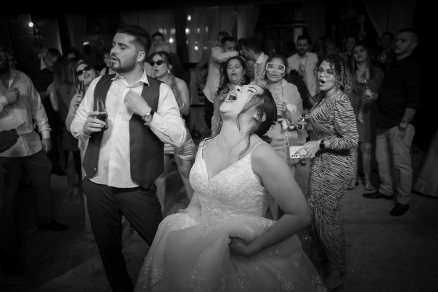 Noiva Gabriela cantando com toda a alma na pista de dança, ao lado de Lucas. Fotografia em preto e branco que transmite a intensidade e a euforia da festa. Momentos de pura liberdade e diversão entre amigos.