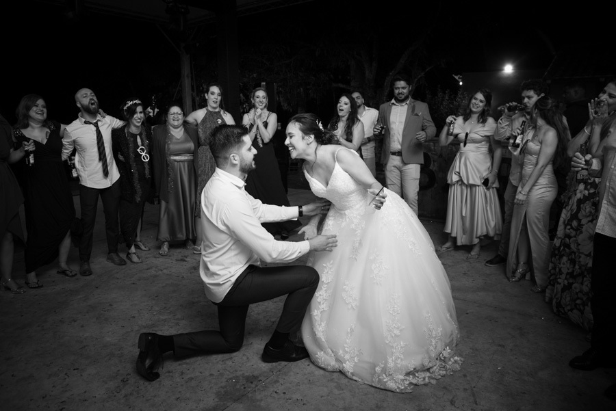 Noivo Lucas se ajoelha na pista para cantar para Gabriela. Uma performance divertida e romântica durante a festa na Quinta das Palmeiras, cercados pelos convidados. O amor celebrado com muita descontração.