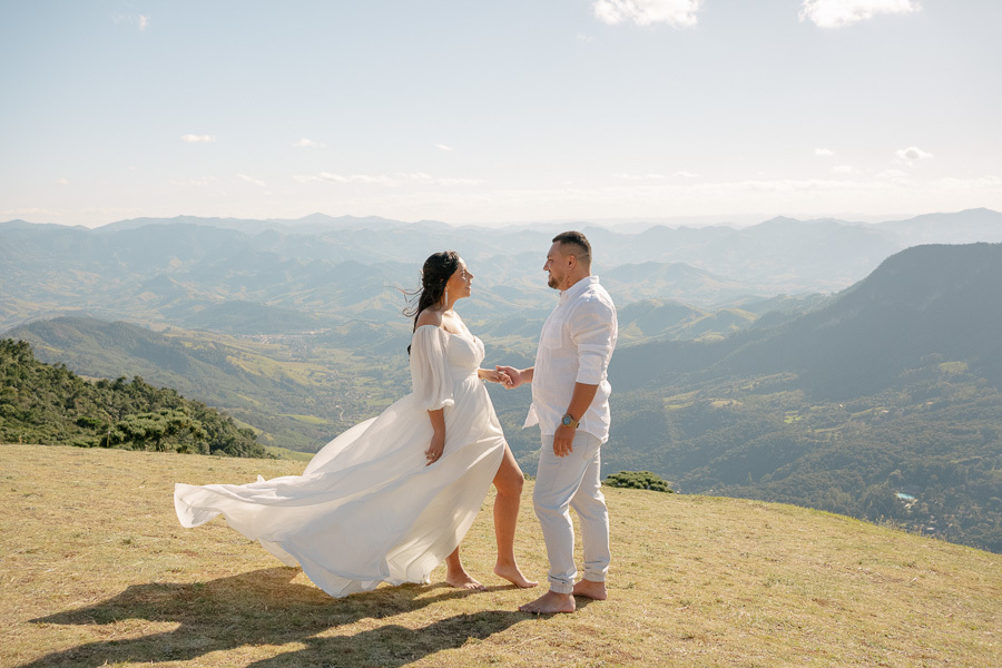 Letícia e Juliano estão de mãos dadas em um campo gramado no topo de uma montanha. Ela veste um vestido branco longo e fluido que esvoaça ao vento; ele usa camisa branca e calça clara. Ao fundo, uma vista panorâmica de vales e montanhas sob um céu azul cl