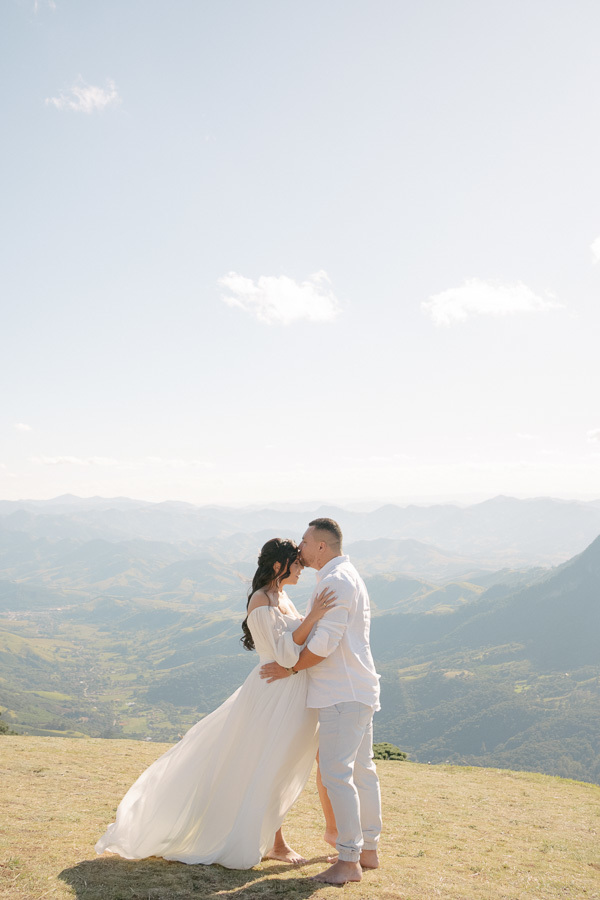 Juliano beija a testa de Letícia no topo da Pedra do Baú em São Bento do Sapucaí. Ela usa um vestido branco longo e fluido que voa levemente; ele veste camisa e calça claras. Ao fundo, uma vasta paisagem de montanhas e vales da Serra da Mantiqueira sob um