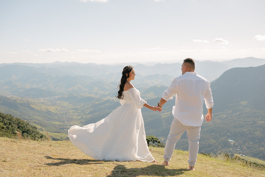 uliano e Letícia estão de mãos dadas no topo da Pedra do Baú, em São Bento do Sapucaí, olhando para o horizonte. Ela veste um vestido branco fluido que esvoaça lateralmente; ele usa camisa branca e calça clara. A imagem captura o casal de costas, destacan