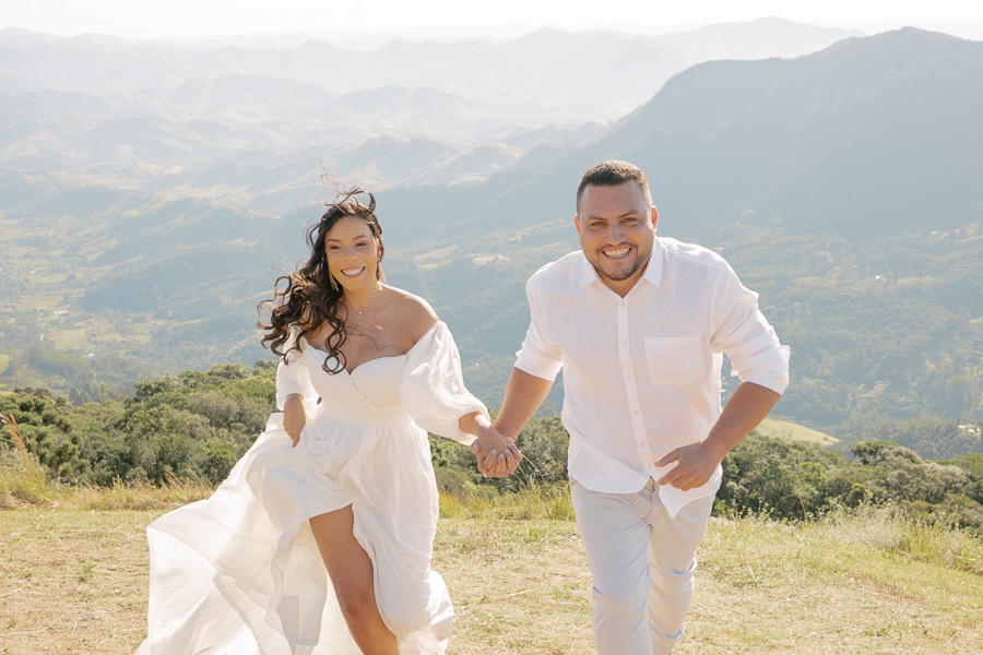 Leticia e Juliano correm sorridentes e de mãos dadas pelo topo gramado da Pedra do Baú. Ela veste um vestido branco fluido com fenda; ele usa camisa branca e calça clara. Ao fundo, a imensidão das montanhas da Serra da Mantiqueira se estende sob a luz 