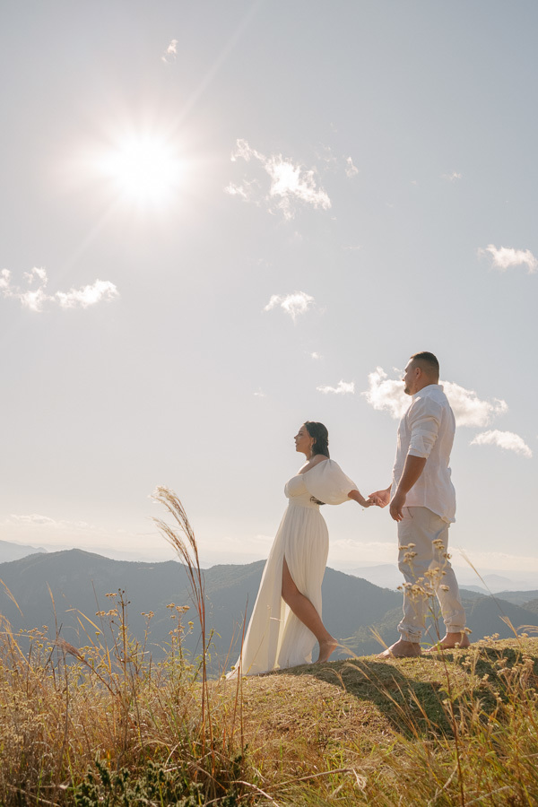 Letícia caminha à frente de Juliano em um campo alto, segurando sua mão. Ela veste um vestido branco com fenda e ele camisa branca. O casal está de perfil contra um sol radiante no céu azul, com montanhas ao fundo e vegetação seca em primeiro plano.